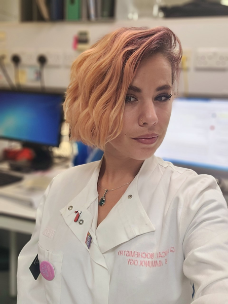 A woman poses in a lab coat in front of a computer screen.