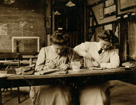 Two women in Victorian dress working at a bench on wood-engraving skills.