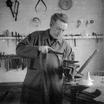 Greyscale photograph of a man, Dunstan Pruden, in silversmithing workshop with hammer and small anvil held in a vice.