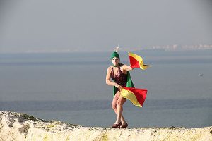 Liz Aggiss dancing on white rock with sea and distant city in background with a green bathing hat and waving yellow and red semaphore flags