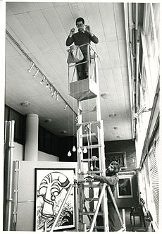Black and white image of art curator on a cherry picker during a picture hanging.
