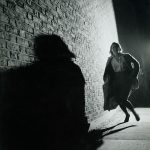 Thurston Hopkins, Woman in trouble, large shadow dominates foreground of picture of woman running towards camera down a high walled street.