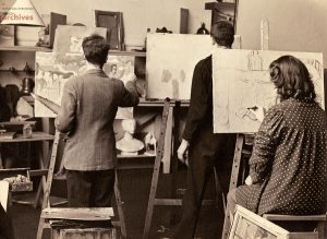 Art studio room with closely packed easels and students, one in a dress another in a tweed jacket, painting