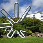 In a garden area, a large tubular matt silver sculpture reflecting the patterns caused by a magnet on an oscilloscope.