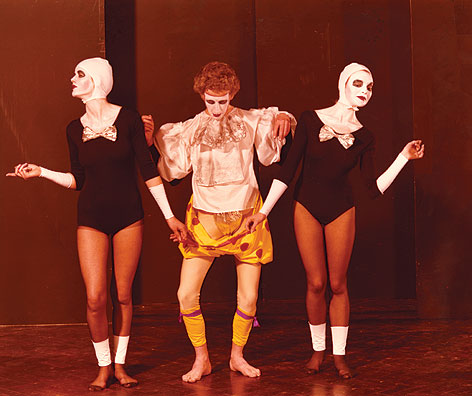 Stylised colour image circa 1948 of three performers in leotards and white masks, central performer in frilled white shirt and yellow spotted shorts.