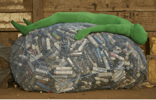 picture of mania a green full body suit laying on a large netted bag full of aerosol cans