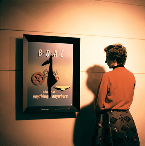 Woman in orange jumper looking at a framed advertising image for BOAC at the British Design Exhibition in North America (no date), picture taken from the Willy de Majo Archive housed at the University of Brighton Design Archives.