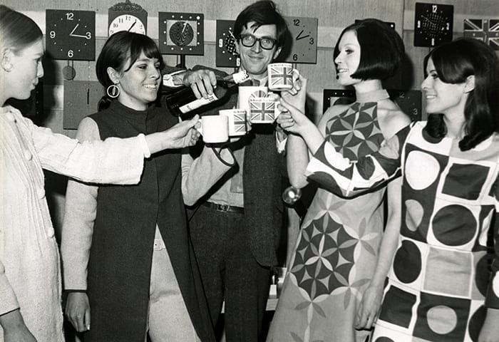 A black and white photograph of Paul Clark making a toast with four women (no date). Taken from the Paul Clark Archive housed at the University of Brighton Design Archives.