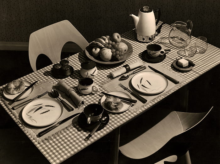 A breakfast table setting for two, which is part of the 'Britain at Table' display in The Design Centre, July - September, 1956. Formica-topped table, two stacking chairs 'Jason’, Irish linen tablemats and napkins. 'Green Wheat' stoneware. Electric coffee percolator. Water jug and glasses. Cutlery and flatware with walnut handles.