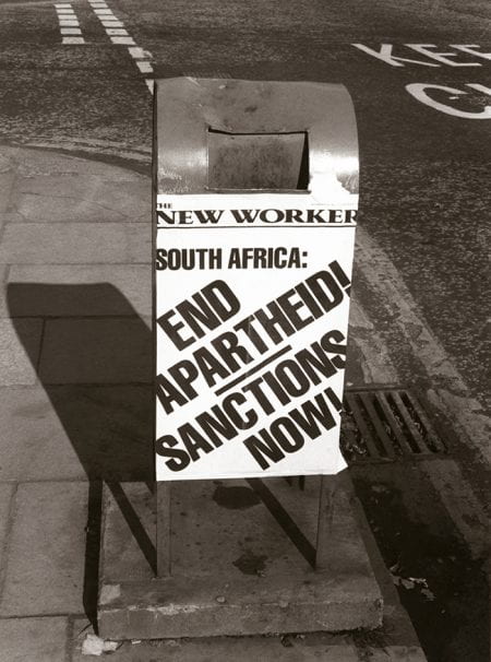 Black and white photo of litter bin with ad for The New Worker - South Africa: End Apartheid! Sanctions Now!