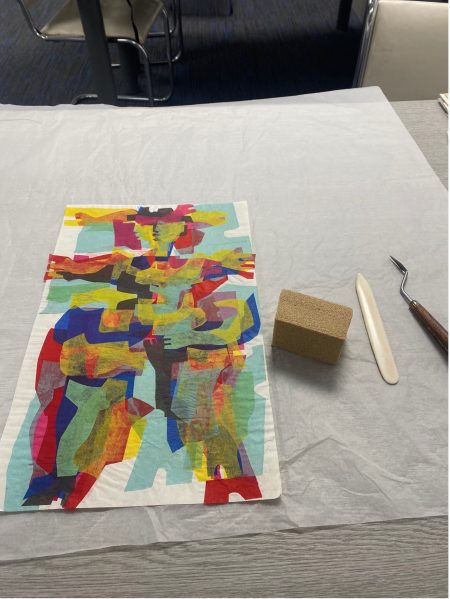 Photograph of a coloured paper collage by Theo Crosby. The images appears to be an abstract representation of a human figure using different geometrical-shaped pieces of coloured paper. The artwork sits on white tissue paper and next to it are several conservation tools used to clean the artwork.