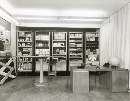 Black and white image showing a designer's office with products on shelves behind a desk