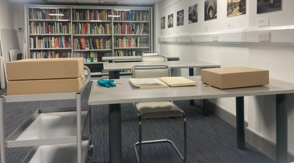 Photograph of the Design Archive's reading room. In the foreground is a desk with files, a pencil, gloves and a brown box on top, a trolly with archive boxes is to the side of the desk.