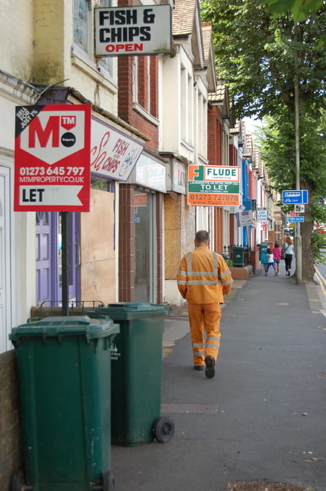 Man in orange overalls walks down a high street pavement with boarded shops and rental signs.