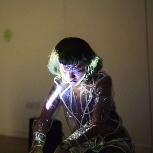 Performance Artist Ada Hao. Projection lit image of artist looking down, dressed in transparent plastic raincoat.