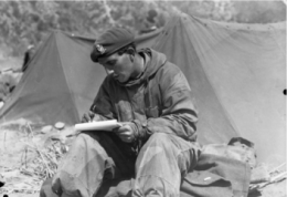 Black and white photograph of man in military clothes and cap writing in a book in front of tents