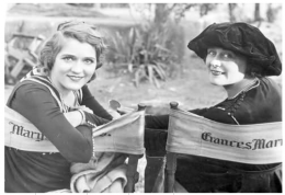 Two women with hairstyles circa 1920 look towards the camera, leaning round on directors style, canvass backed chairs with visible names Mary and Frances in Gothic lettering, surnames obscured.