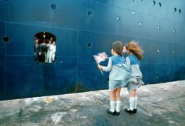 Two young girls in identical blue dresses stand on a dockside with Union Jack flags while a large ship departs, figures visible in the entranceway.