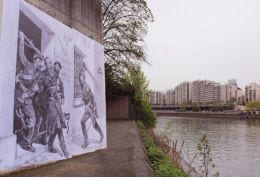 Large picture on a river front, detailed drawing of military activity visible. Distant bank of wide river has blocks of high-rise flats.