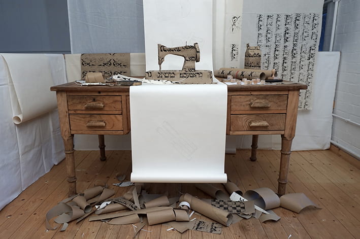 An art installation featuring a cardboard sewing machine with Hebrew writing on it on a sewing table.