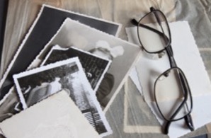 A black and white image of some photos on a desk with a pair of reading glasses on top.