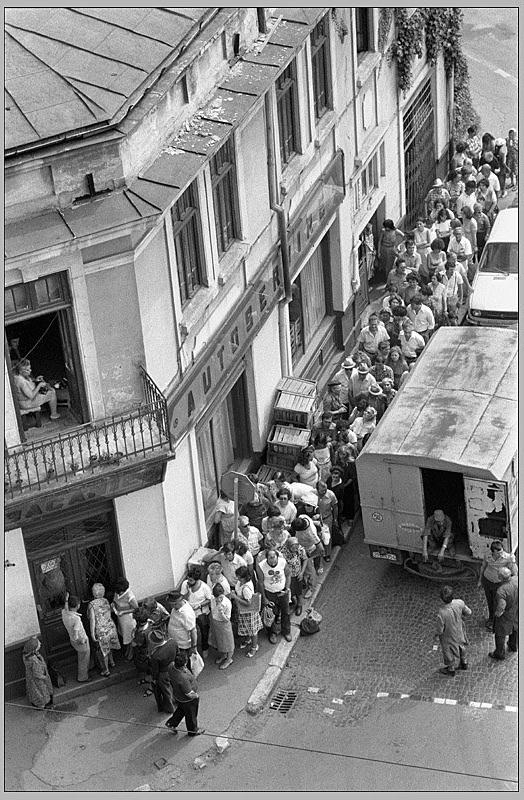 A black and white photo taken from above looking down on a queue of people waiting to enter a shop on the street in Romania 1982.