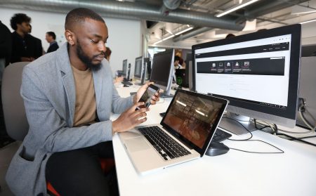 Male student looking at app on phone siting in front of laptop and desktop computers