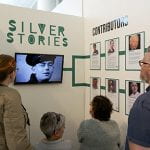 Exhibition booth with title Silver Stories on a wall, a monitor playing films and foamboards with information on project and participants in digital storytelling