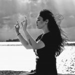 Black and white photo of a woman in a yoga pose, hands raised, hair blowing in the breeze. In the background is a sunlit lake.