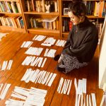 A woman sits on a wooden floor, gazing contemplatively at numerous slips of paper laid out before her.. In the background are bookshelves.