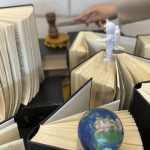 Two small objects stand on the pages of upright books: an astronaut and a world globe. In the background, a hand bangs a gavel.