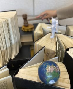 Two small objects stand on the pages of upright books: an astronaut and a world globe. In the background, a hand bangs a gavel.