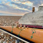 Photo of an upturned canoe and boat on a shingle beach. Four Russian dolls stand on the canoe in descending height. On the boat stands one large Russian doll.