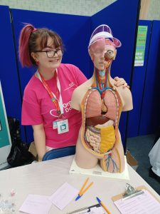 Estella with pink hair wearing a pink NHSBT t-shirt posing with the torso which is wearing a pink visor.