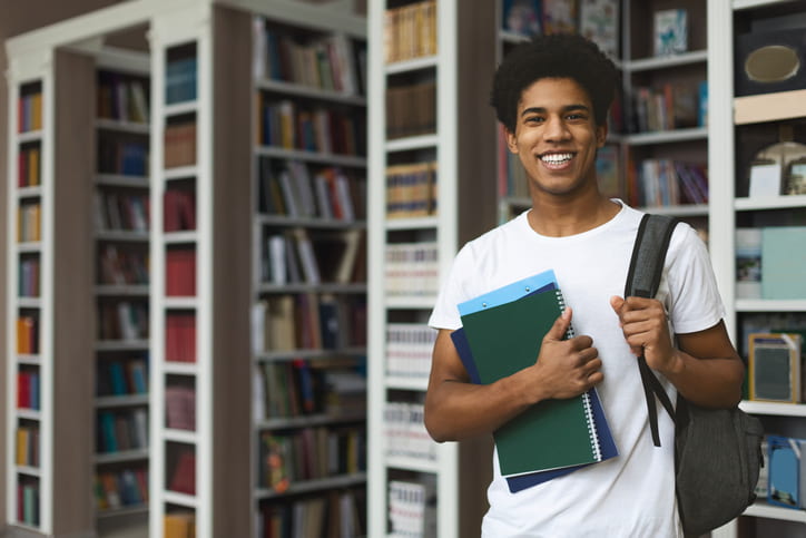 Smiling Student with folders stood in front of bookshelf at uni