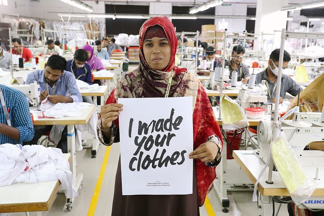 Image of a woman with a red head scarf holding a sign that says "I made your clothes." She is in a factory. 