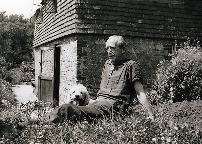 Black and white photo of Max Gill sitting in a cottage garden with a white dog