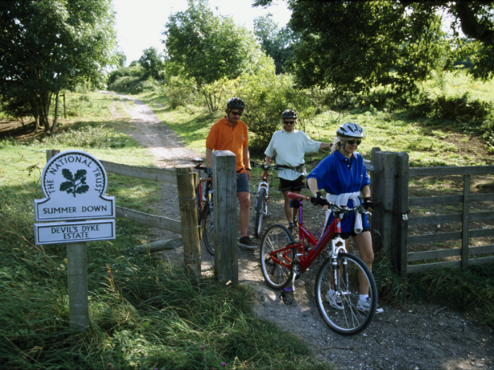 Three cyclists come through a gate on a pathway somewhere in the South Downs countryside. (Source: Brighton and Hove City Council, 2022)