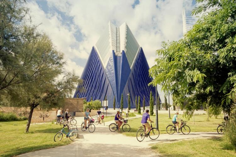A large building sits in the middle of the frame surrounded by a green park with many cyclists. (Source: Visit Valencia 2022)