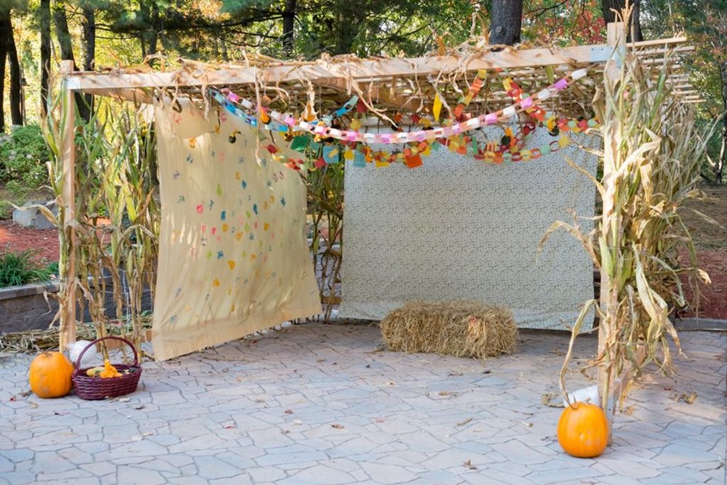 Three sided temporary structure with a wooden frame, draped material for walls, paper bunting and a bale of hay as a seat inside it. Tall corn stalks are woven into the walls.