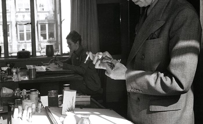 Black and white portrait of Reco Capey inspecting a 3D item with a woman sitting at a desk in the background