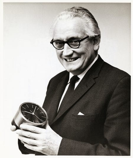 Black and white portrait of Jack Howe smiling at the camera holding a desk clock