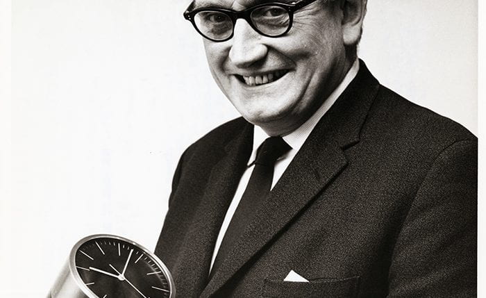 Black and white portrait of Jack Howe smiling at the camera holding a desk clock