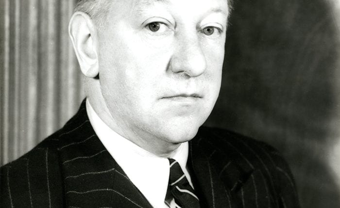 Black and white portrait of Allan Walton in a striped suit and tie posed sideways looking into the camera.