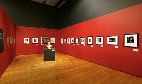 Gallery space with red walls and polished wooden floor. Closely hung line of pictures along the two visible walls