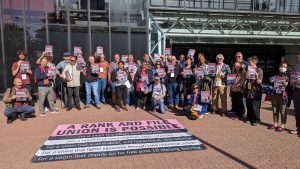 Large group of people standing outside UCU Congress 2023 venue in the sunshine, holding posters saying Save Brighton University