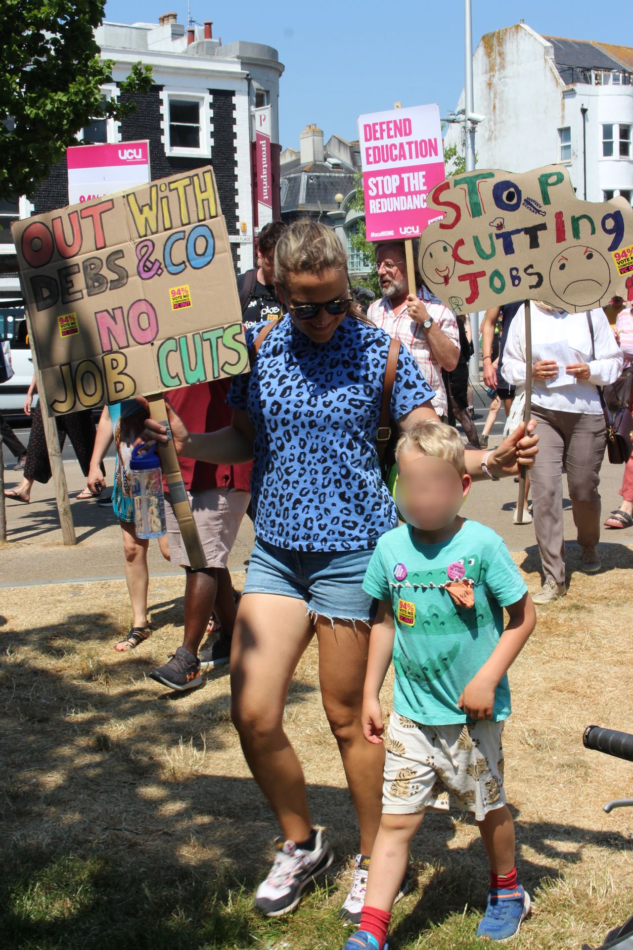 Mum and young son at march. Mum holding placard "Out with Debs & co. No job cuts"