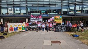 Several dozen people standing outside Checkland Building, Falmer campus. Holding placards and banners including Yellow banner "94% vote no confidence. Vice Chancellor Debra Humphris Out". Black banner "Save Brighton University. Stop the Redundancies"